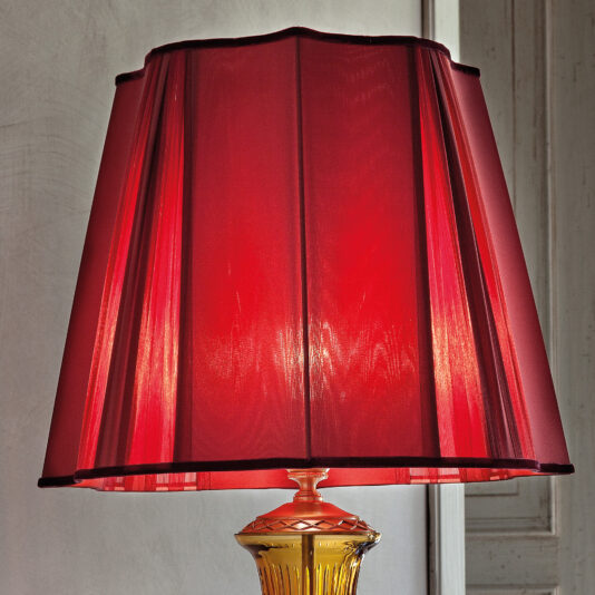 Classic Amber Glass Table Lamp With Ruby Stones A close-up of the Classic Amber Glass Table Lamp With Ruby Stones, featuring a red lampshade with a pleated design and even vertical lines. The lampshade is accented with dark trim at the top and bottom. In the background, a softly lit, neutral-colored wall adds to the lamp's elegant appearance.