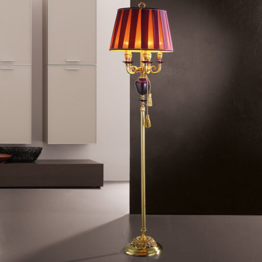 A stylish Classic Candelabra Style Speckled Glass Floor Lamp with a gold base stands in front of a dark wall. It features three candle-like bulbs, reminiscent of a classic candelabra style, and a decorative black and red striped lampshade. In the background, modern minimalistic furniture with clean lines completes the elegant setting.
