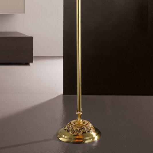 A close-up shot highlights the base of the Classic Candelabra Style Speckled Glass Floor Lamp, showcasing its ornate detailing and elegant brass finish. The background features a white wall and dark wood shelf, creating a striking contrast with the lamp's golden color and speckled glass accents.