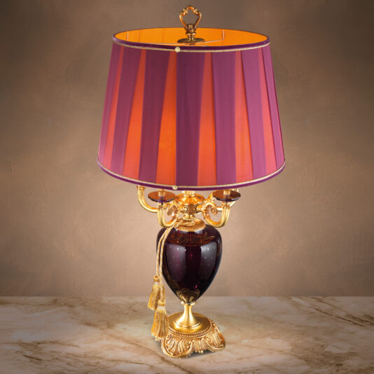 The Classic Candelabra Style Speckled Glass Table Lamp features intricate gold detailing and a deep maroon base. Its maroon lampshade, complete with a golden inner lining, creates a warm glow. Accentuated with a gold tassel hanging from the base, this lamp sits elegantly on a speckled glass marble surface.