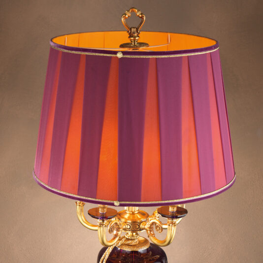 The Classic Candelabra Style Speckled Glass Table Lamp boasts a golden base with a luxurious purple lampshade adorned with a subtle vertical stripe pattern. Its golden interior casts a warm, inviting glow. The ornate design is enhanced by gold-toned detailing and an elegant top finial, evoking candelabra-style sophistication.