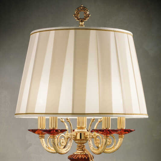 The Classic Candelabra Style Table Lamp boasts a luxurious cream-colored silk lampshade with a wide design. Its ornate brass base is adorned with intricate scrollwork and decorative red glass elements, adding an elegant touch. The lamp features multiple candle-style bulbs and is topped off with a decorative finial.