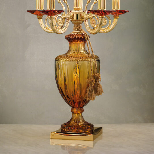 A detailed close-up of the Classic Candelabra Style Table Lamp reveals ornate designs in gold and amber glass with multiple candle holders. The base is embellished with a gold rope tassel, and this elegant piece is set on a reflective marble surface against a muted, textured background.