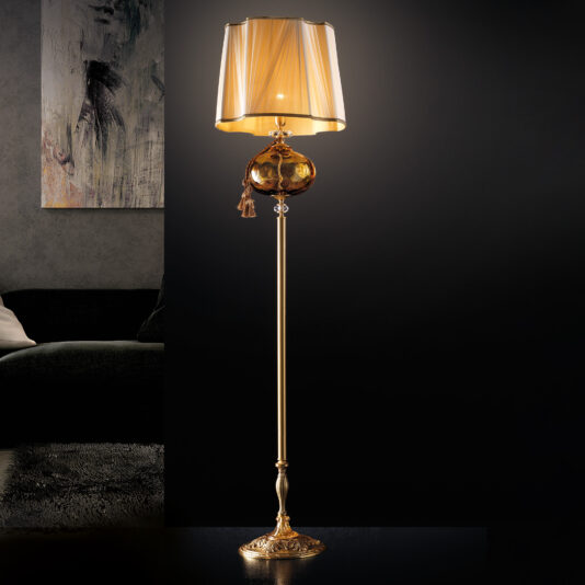 A tall, elegant floor lamp with a golden metallic stand and an ornate base, the Classic Dimpled Amber Glass Floor Lamp features a spherical decorative element beneath its translucent, pleated golden lampshade. It stands against a dark background next to a framed abstract artwork and a dark sofa.