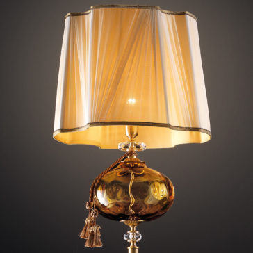 Classic Dimpled Amber Glass Floor Lamp A close-up of an ornate table lamp showcases its gold-toned base and decorative, round body adorned with intricate carvings. The beige lampshade, reminiscent of the Classic Dimpled Amber Glass Floor Lamp, has a pleated texture, is trimmed with gold accents, and features a tasseled rope elegantly draped around the base.