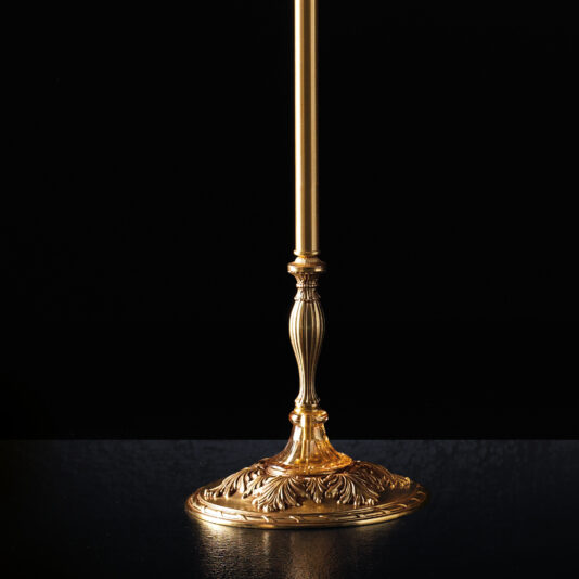 A close-up of an ornate gold-colored lamp base set against a black background, showcasing intricate floral and leaf patterns with a slender, fluted stem extending upward. The design is reminiscent of the Classic Dimpled Amber Glass Floor Lamp. The reflective surface enhances the elegant details of the lamp.