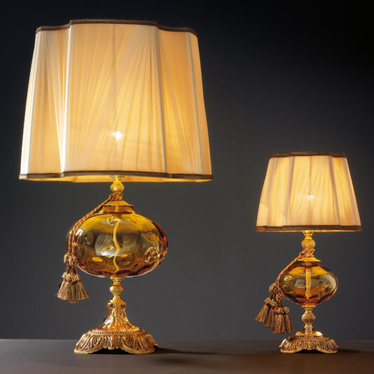 Two ornate table lamps with gold accents and beige fabric shades. The larger lamp on the left is the Classic Dimpled Amber Glass Table Lamp, featuring a classic dimpled amber glass base adorned with intricate tassels, complemented by the smaller one on the right. Both are set against a dark background, exuding timeless elegance.