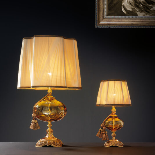 Classic Dimpled Amber Glass Table Lamp Two ornately designed Classic Dimpled Amber Glass Table Lamps, featuring golden bases and spherical accents, are displayed against a dark background. Each lamp is adorned with a pleated cream-colored shade. The taller lamp is positioned on the left, while a framed section of artwork is partially visible in the top right corner.