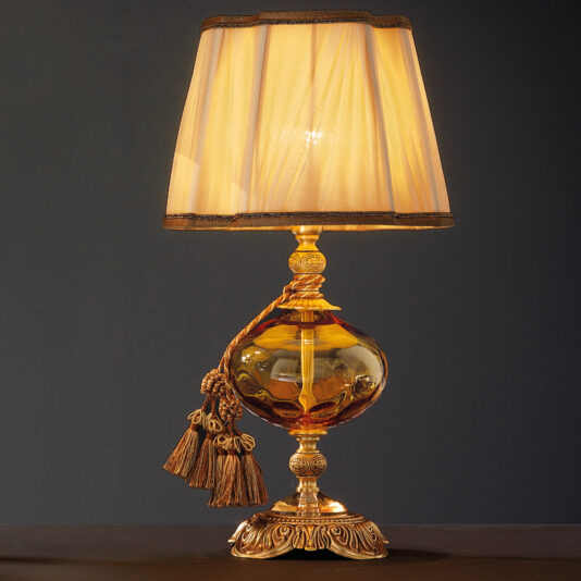 Classic Dimpled Amber Glass Table Lamp The Classic Dimpled Amber Glass Table Lamp boasts a classic dimpled amber glass design paired with an ornate gold base. It is adorned with a pleated, sheer beige shade and decorative gold tassels hanging from the neck. When lit, it casts a warm glow against a dark background.