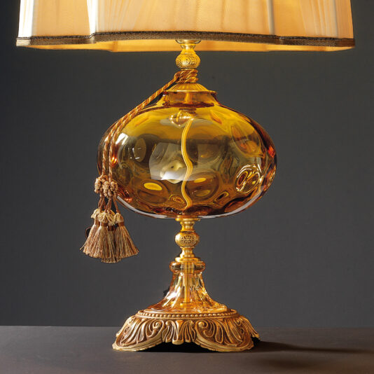 Classic Dimpled Amber Glass Table Lamp A Classic Dimpled Amber Glass Table Lamp featuring a pleated beige lampshade, an ornate golden base, and a decorative amber glass body with intricate designs. A tassel hangs from the neck of the lamp, adding an elegant touch to its vintage aesthetic. The lamp is set against a dark background.
