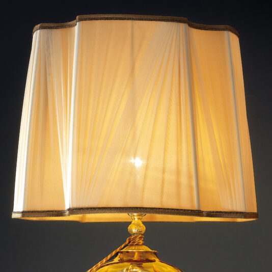 Classic Dimpled Amber Glass Table Lamp A close-up image of the Classic Dimpled Amber Glass Table Lamp showcasing its cream-colored, pleated lampshade and ornate golden base. The lampshade softly diffuses the warm light, casting a gentle glow. The dark background enhances the lamp's warm illumination.