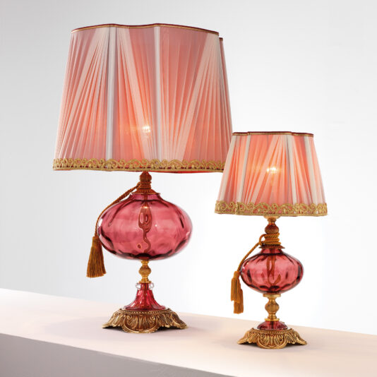 The Classic Dimpled Rose Glass Table Lamp features an elegant design with a dimpled rose glass base and a matching pleated red lampshade adorned with gold trim. Each lamp showcases intricate detailing with gold accents and decorative tassels hanging from the neck. The larger lamp is situated on the left, while a smaller one sits to the right.