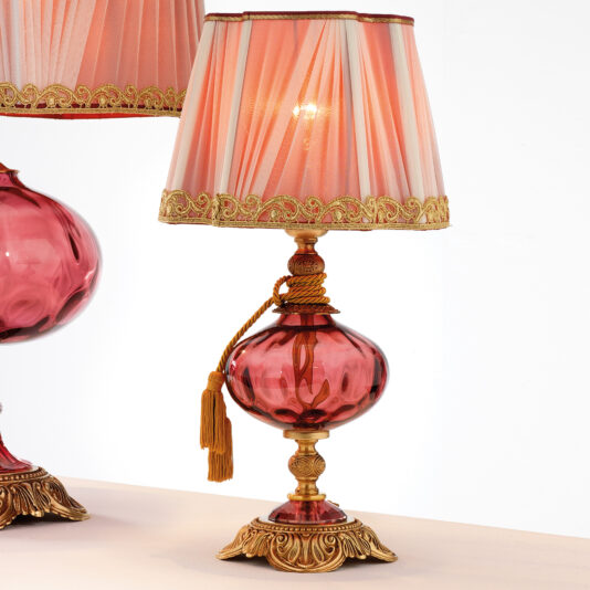 The Classic Dimpled Rose Glass Table Lamp is a decorative piece with a red glass base, adorned with ornate gold accents and a tassel hanging from the neck. It features a pink lampshade with vertical pleats and intricate gold trim. Another lamp of the same design is partially visible on the left side.