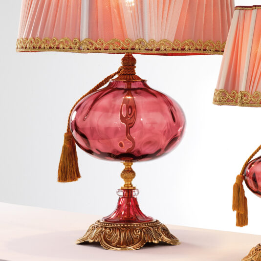 The Classic Dimpled Rose Glass Table Lamp features a pink, ornate base with intricate gold accents, a tasseled pull cord, and a fringed, pink silk lampshade trimmed with gold detailing. Another similar lamp is partially visible beside it.