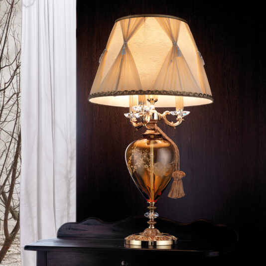 The Classic Engraved Amber Glass Table Lamp, with a beige fabric shade adorned with tasseled details, stands on a dark wooden surface. Its ornate, intricately-designed base features floral patterns. In the background, a dark wall and a small glimpse of a window on the left can be seen.