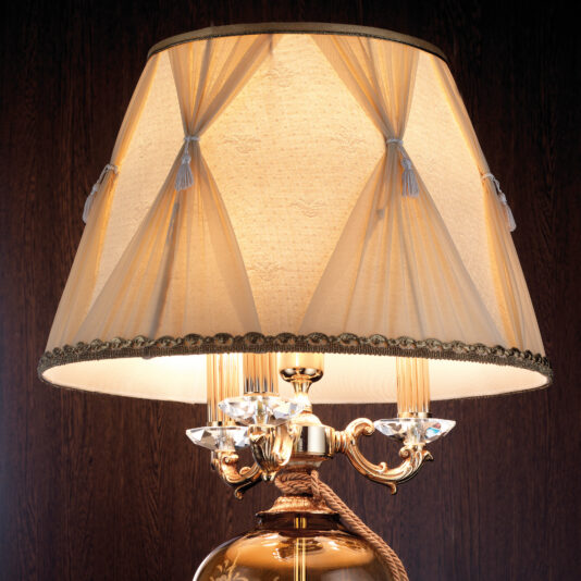Classic Engraved Amber Glass Table Lamp A close-up of the Classic Engraved Amber Glass Table Lamp featuring a beige fabric shade adorned with gold trim and tassels. The lamp stem showcases intricate gold detailing and clear crystal accents. The elegant design of the lamp is highlighted by a dark wooden surface in the background.
