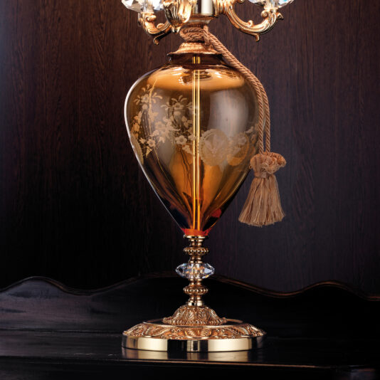 Classic Engraved Amber Glass Table Lamp A close-up of the Classic Engraved Amber Glass Table Lamp, showcasing its transparent amber-colored body with floral etchings and delicate metallic accents. The lamp boasts a gold base and a decorative gold tassel hanging from the top, all beautifully contrasted against a dark wood background.