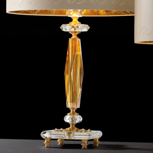 Classic Glass Table Lamp Introducing the Classic Glass Table Lamp, featuring an exquisite gold and crystal base adorned with intricate, faceted crystal sections and elegant gold detailing. This sophisticated piece is topped with a beige lampshade, creating a polished look when positioned against a striking black background.