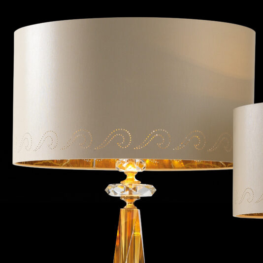 Classic Glass Table Lamp The close-up highlights the Classic Glass Table Lamp, showcasing two pieces adorned with beige, cylindrical lampshades featuring a delicate perforated swirl design along the bottom edge. The transparent, golden bases resemble crystal and boast an intricate geometric shape.