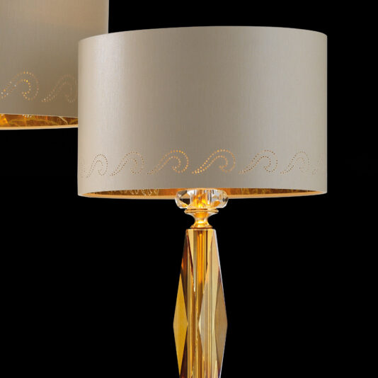 Classic Glass Table Lamp Introducing the Classic Glass Table Lamp: Featuring a beige drum shade adorned with a wavy dotted pattern, this elegant lamp boasts a golden faceted base. The gold and beige color scheme is stunningly highlighted against the black background, enhancing its sophisticated design.