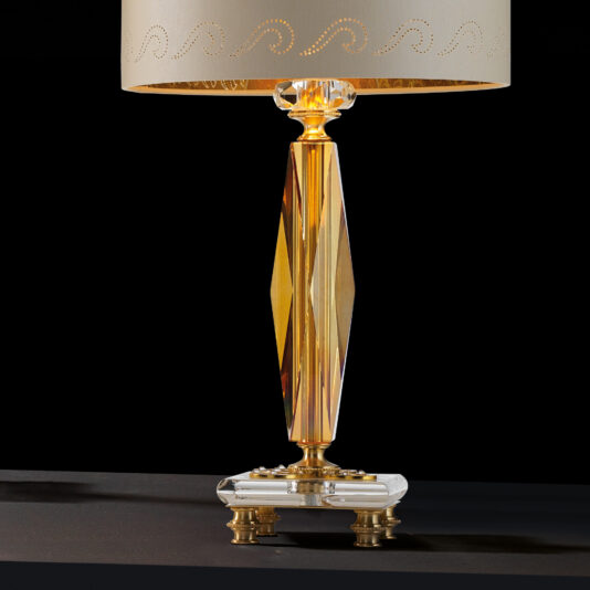 Classic Glass Table Lamp The Classic Glass Table Lamp, showcasing a white perforated fabric shade, a geometric, faceted glass base accented with gold metal, casts a warm light as it rests on a dark surface against a black background.