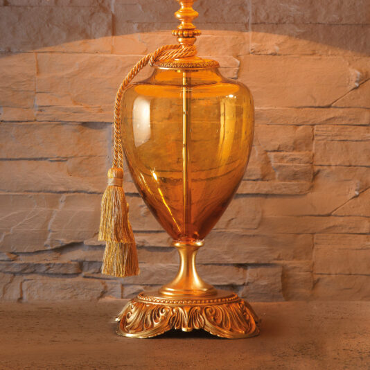A stunning Classic Speckled Amber Glass Table Lamp with a gilded base and lid is displayed against a stone wall. The lamp, adorned with a gold decorative tassel hanging from the top, exudes an ornate and timeless elegance.