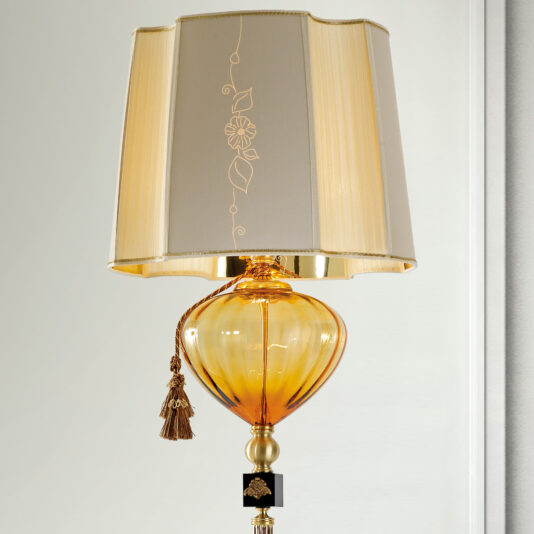 Classic Style Amber Glass Floor Lamp A close-up of the Classic Style Amber Glass Floor Lamp showcases its golden, intricately designed base with a heart-shaped amber glass bottom. The beige lampshade, adorned with floral embroidery and a decorative tassel, enhances the classic style against a plain, light gray wall.