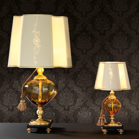 Two Classic Style Amber Glass Table Lamps with gold bases and intricate designs exhibit a timeless elegance. Positioned on a dark surface against a textured backdrop, each lamp showcases a white lampshade embellished with decorative floral patterns and a tassel hanging from the base, accented by amber glass details.