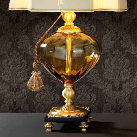 Classic Style Amber Glass Table Lamp Introducing the Classic Style Amber Glass Table Lamp, featuring a sophisticated design with a large amber-colored glass base and intricate gold metal accents. The lamp is adorned with a rectangular beige lampshade and complemented by a decorative tassel hanging from the shade. It stands on a black base with small gold feet, all set against a textured dark wallpaper backdrop.