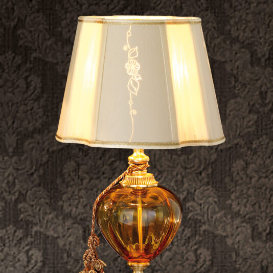 Classic Style Amber Glass Table Lamp The Classic Style Amber Glass Table Lamp features an amber glass base and a cream-colored fabric shade adorned with a delicate floral pattern. It emits a warm, inviting light and includes a decorative tassel hanging from the base, making it perfect against textured dark wallpaper.