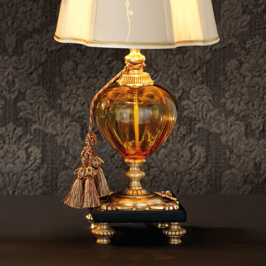 Classic Style Amber Glass Table Lamp The Classic Style Amber Glass Table Lamp features an ornately designed amber glass base with gold accents, paired with a beige lampshade. Its sophisticated charm is heightened by intricate details and a braided tassel adorning the neck. The lamp sits elegantly on a black surface against a textured, dark background.