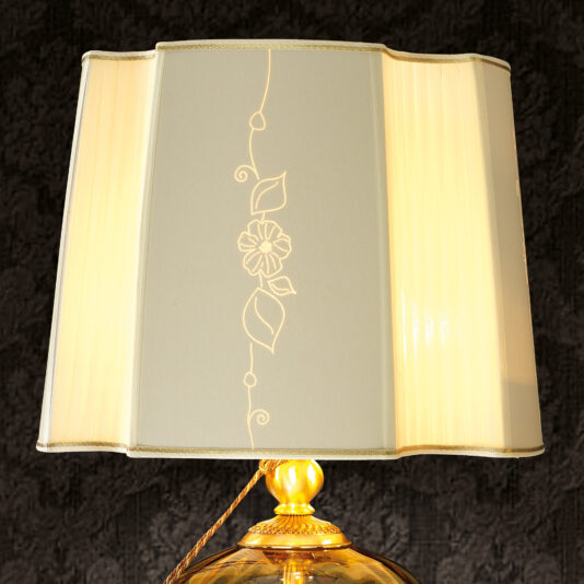 Classic Style Amber Glass Table Lamp A Classic Style Amber Glass Table Lamp with a rectangular beige fabric lampshade that showcases an intricate embroidered floral design. The ornately designed base is crafted from amber glass, accented with golden details. When turned on, the lamp emits a warm glow against a dark, textured background.