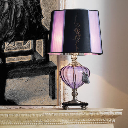 A Classic Style Violet Glass Table Lamp with a dual-colored lampshade (purple and black) stands on a beige mantel. The background features a monochrome portrait, partially visible. The lamp has a small decorative tassel hanging from its neck.