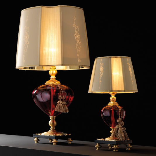 A pair of the Classic Style Ruby Glass Table Lamps elegantly stands on a black surface against a dark background. The lamps feature gold-trimmed, cream-colored shades and bases made of ruby glass with gold accents and decorative tassels. One lamp is noticeably taller than the other, adding a unique touch to their classic design.
