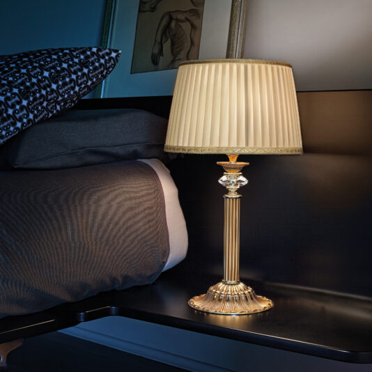 A stylish Classic Table Lamp With Swarovski Detail, featuring a golden base and a pleated shade, is illuminated on a dark wooden nightstand next to a bed with dark bedding. In the background, partially visible framed artwork enhances the room’s sophisticated ambiance.