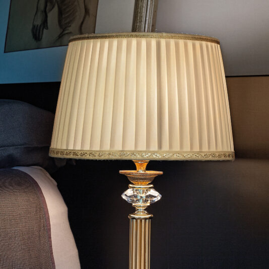 A close-up shot of the Classic Table Lamp With Swarovski Detail, featuring a pleated beige lampshade and a glass crystal base. The lamp is placed on a bedside table next to a gray pillow. A framed artwork is partially visible on the wall behind the lamp.