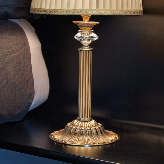 The Classic Table Lamp With Swarovski Detail, featuring an elegant golden finish, a fluted design, and an ornate base adorned with Swarovski crystals, stands on a black nightstand next to the bed. Its beige lampshade with a subtle pattern perfectly complements the luxurious, classic decor of the bedside area.