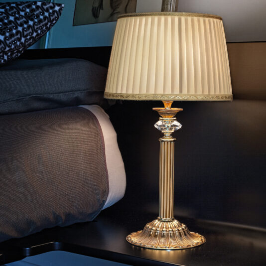 A Classic Table Lamp With Swarovski Detail and a pleated beige shade stands next to a neatly made bed with gray and white bedding. The lamp is turned on, casting a warm, inviting glow in the dimly lit room.