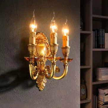 A Triple Candle Style Wall Light with an ornate, vintage design and gold finish illuminates the textured wall with its three flame-shaped bulbs, casting a warm glow. The elegant sconce, adorned with intricate detailing, is mounted next to a bookshelf brimming with books and decorative items.