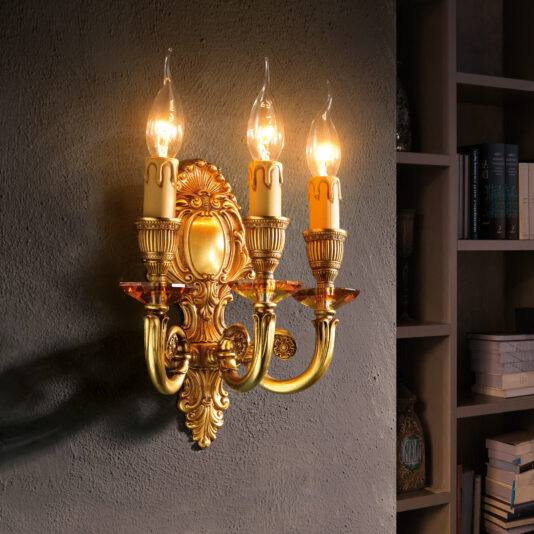 A Triple Candle Style Wall Light with an ornate, vintage design and gold finish illuminates the textured wall with its three flame-shaped bulbs, casting a warm glow. The elegant sconce, adorned with intricate detailing, is mounted next to a bookshelf brimming with books and decorative items.