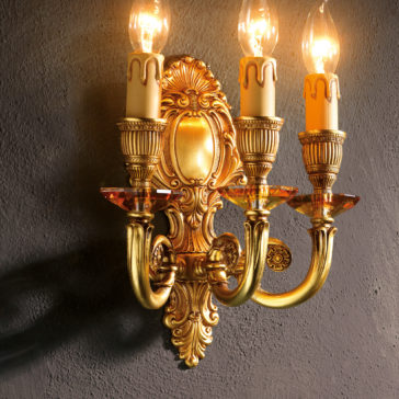 The elegance of the Triple Candle Style Wall Light is embodied in this luxurious, ornate gold wall sconce. Featuring intricate detailing and three candle-like bulbs, it casts a warm light against a textured dark gray wall.