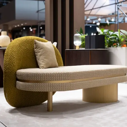 Modern beige chaise longue with a textured olive green backrest, placed in a well-lit showroom. Showcasing the essence of luxury hallway furniture, this piece features a small cushion resting on the seat; nearby are plants, a water glass, and shelves, all complementing the contemporary decor theme.