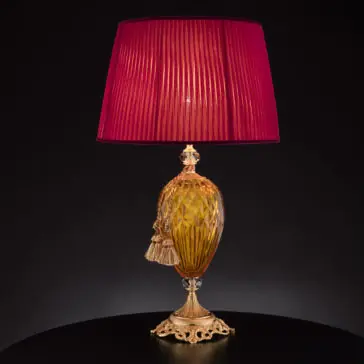 Diamond Faceted Amber Glass Table Lamp Introducing the **Diamond Faceted Amber Glass Table Lamp**: This elegant table lamp features an ornate golden base and body adorned with intricate patterns and diamond-faceted amber glass accents. The vibrant red pleated fabric lampshade, set against a black background, enhances its sophisticated design. Two decorative tassels hanging from the neck add a final touch of refinement.