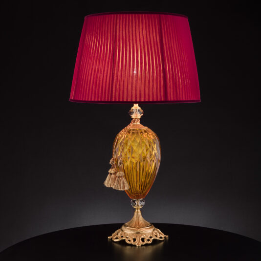 Introducing the **Diamond Faceted Amber Glass Table Lamp**: This elegant table lamp features an ornate golden base and body adorned with intricate patterns and diamond-faceted amber glass accents. The vibrant red pleated fabric lampshade, set against a black background, enhances its sophisticated design. Two decorative tassels hanging from the neck add a final touch of refinement.