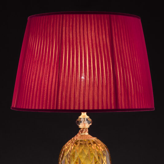 The Diamond Faceted Amber Glass Table Lamp features a pleated, vibrant red lampshade. Its base is intricately designed with crystal-like geometric patterns and boasts a golden hue. A solid black background accentuates the lamp's striking design and vivid colors.