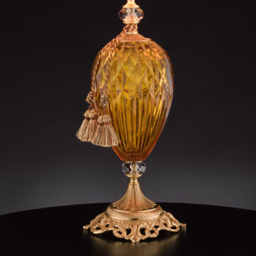 Diamond Faceted Amber Glass Table Lamp The Diamond Faceted Amber Glass Table Lamp, adorned with intricate carvings and showcasing a textured surface, is elegantly displayed against a black background. It features gold accents and a pair of tassels draped from the top, all mounted on an ornate gold pedestal reminiscent of an elegant table lamp.