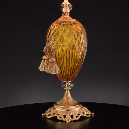 The Diamond Faceted Amber Glass Table Lamp, adorned with intricate carvings and showcasing a textured surface, is elegantly displayed against a black background. It features gold accents and a pair of tassels draped from the top, all mounted on an ornate gold pedestal reminiscent of an elegant table lamp.