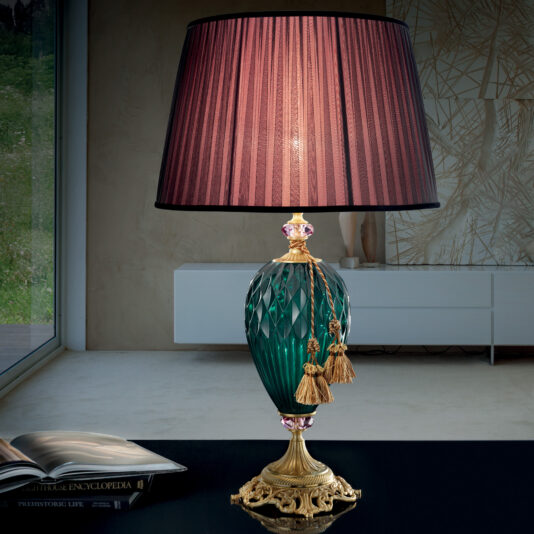 The Diamond Faceted Green Glass Table Lamp, featuring an ornate gold base and a dark pink pleated lampshade adorned with gold tassels, is gracefully placed on the table. Next to the lamp lies an open book.