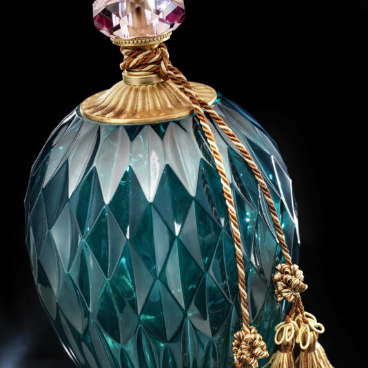 Diamond Faceted Green Glass Table Lamp A luxurious Diamond Faceted Green Glass Table Lamp featuring a gold-colored ornate neck and a decorative stopper. The lamp is adorned with golden braided cords and tassels, set against a dark background, highlighting its elegance and intricate craftsmanship.