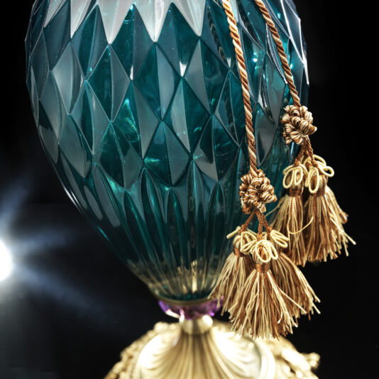 Diamond Faceted Green Glass Table Lamp A close-up of the Diamond Faceted Green Glass Table Lamp, showcasing its ornate design. The lamp features a textured, diamond-patterned green surface with golden tassels hanging from it, and an intricately designed base with gold detailing. The dark background makes the lamp stand out beautifully.